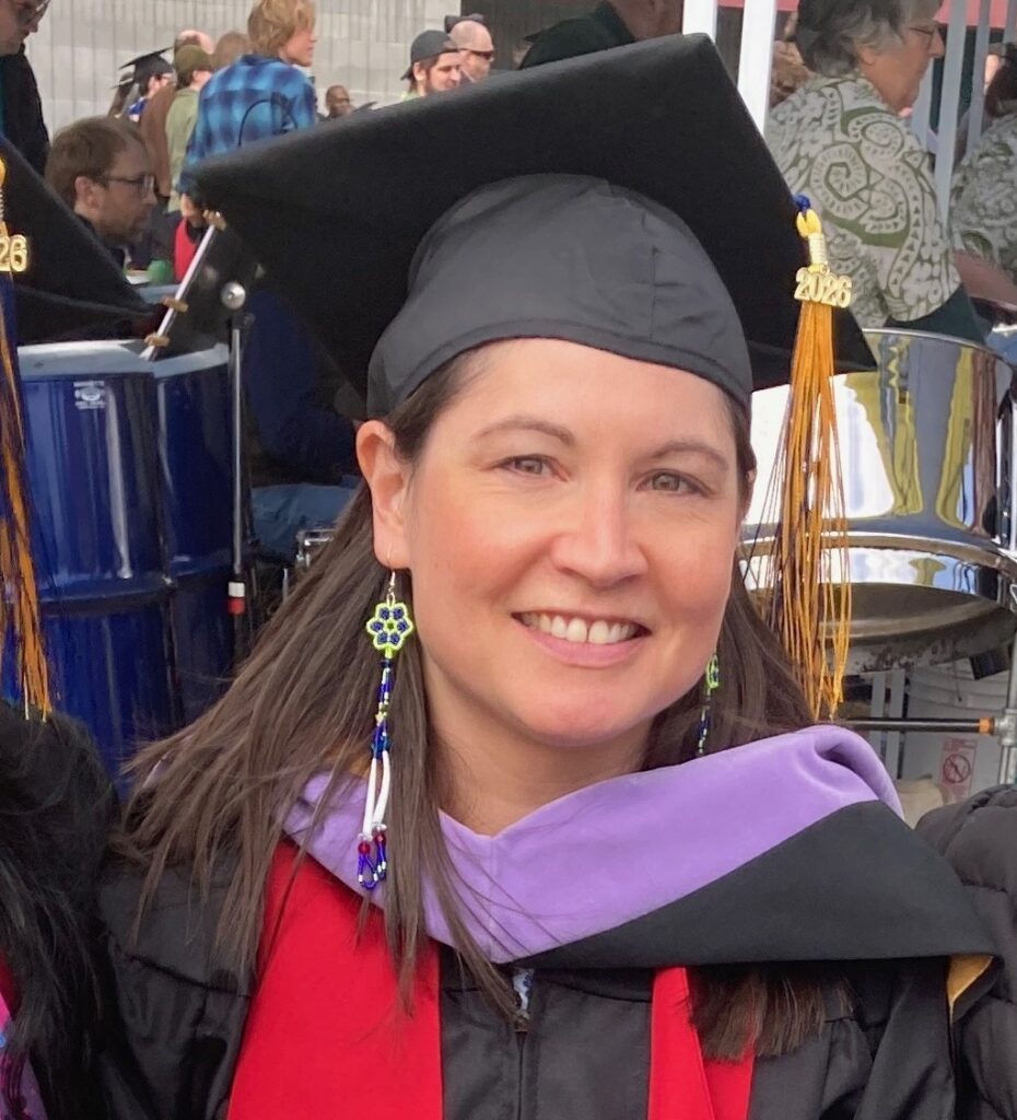 Author Dana Diehl smiling at her 2026 graduation ceremony, wearing a black mortarboard with a gold '2026' tassel, black graduation gown, lavender master's hood, red Alaska Native heritage sash, and beaded earrings. A drum kit and crowd are visible in the background.